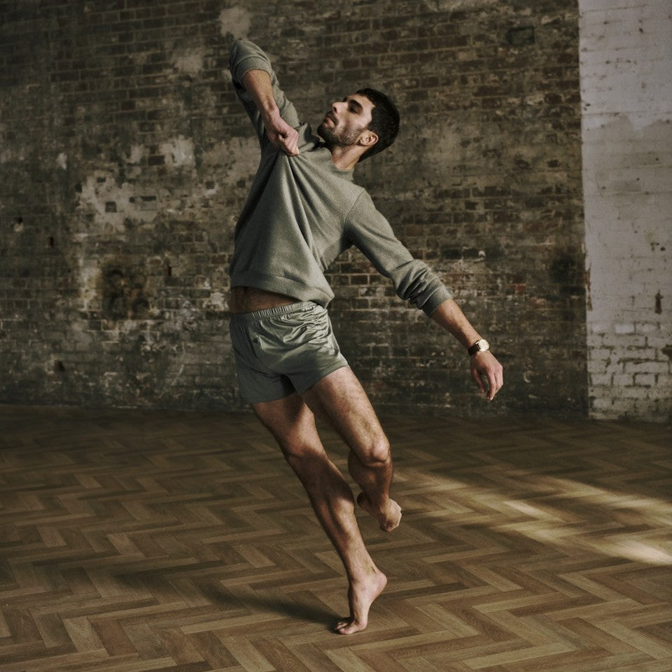 Man wearing HANRO Cotton Essentials black tank top and khaki boxer shorts, mid-jump against exposed brick wall, AW26 campaign